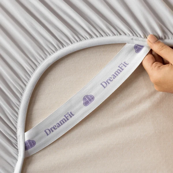 Hand lifts sheet edge to reveal a white DreamFit elastic band printed with purple logos.