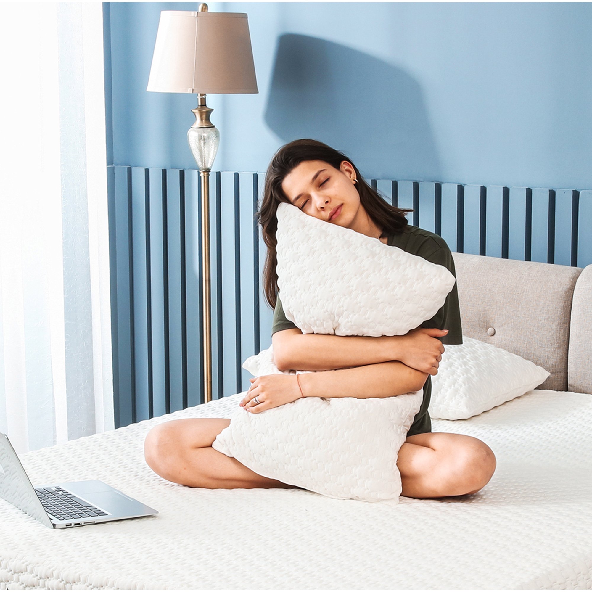 Woman sitting cross-legged on a bed, eyes closed as she hugs and rests on a textured white pillow.